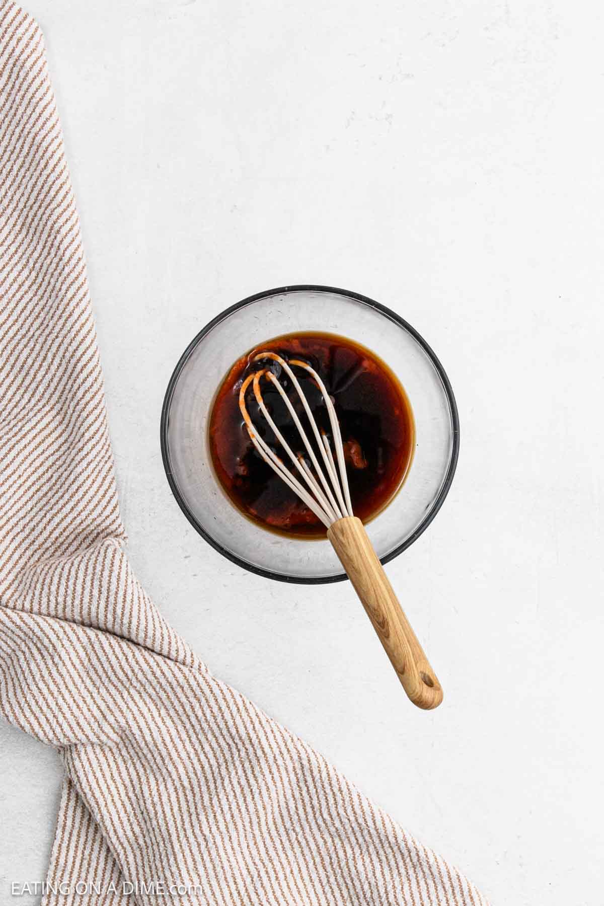 A glass bowl filled with dark Chicken Ramen sauce and a wooden-handled whisk, placed on a light surface next to a beige and white striped cloth.
