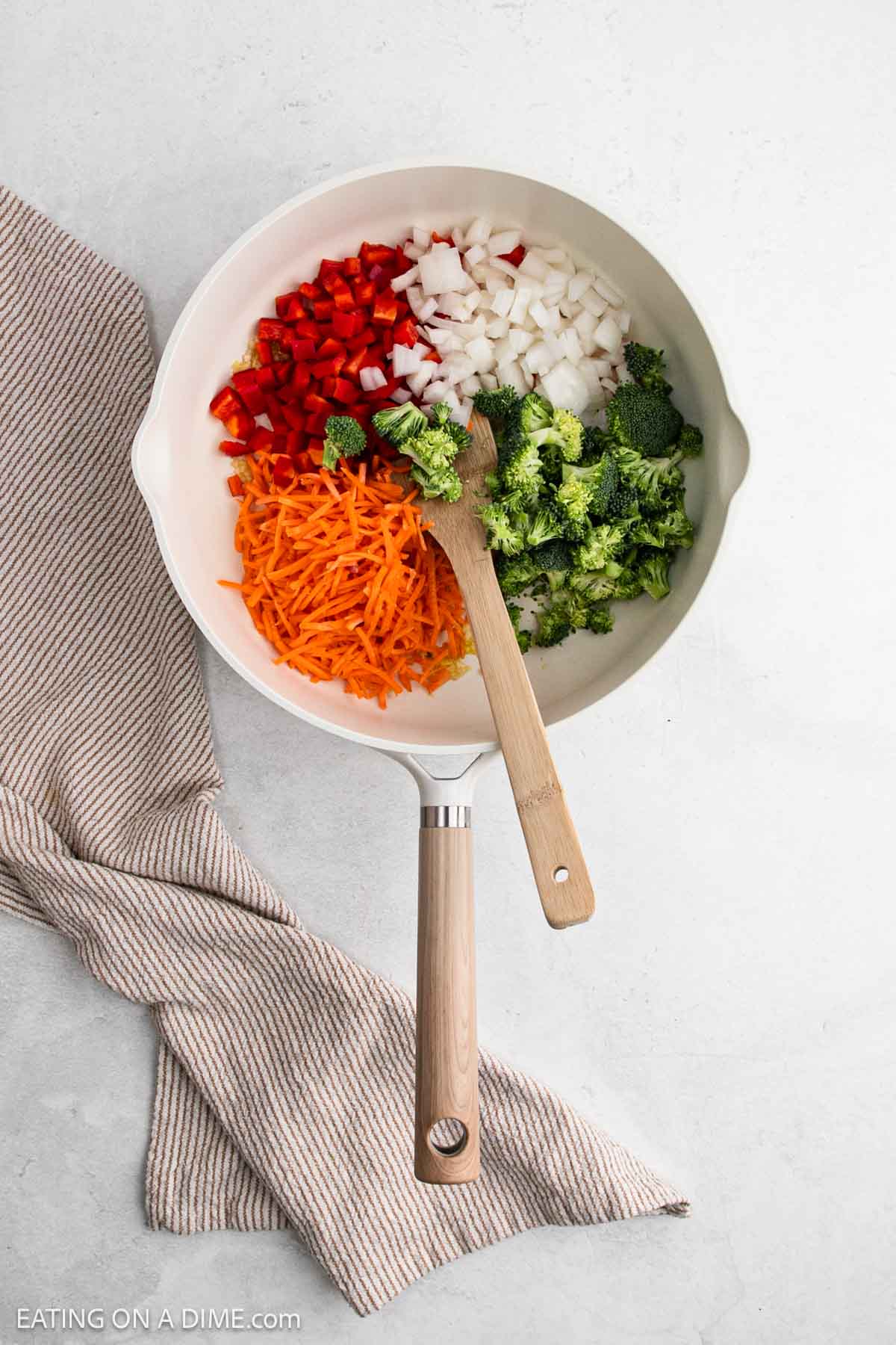 Overhead view of a white skillet containing Chicken Ramen ingredients—chopped red bell pepper, diced onion, shredded carrot, and chopped broccoli—with a wooden spoon resting inside. The skillet is on a light surface with a striped cloth nearby.