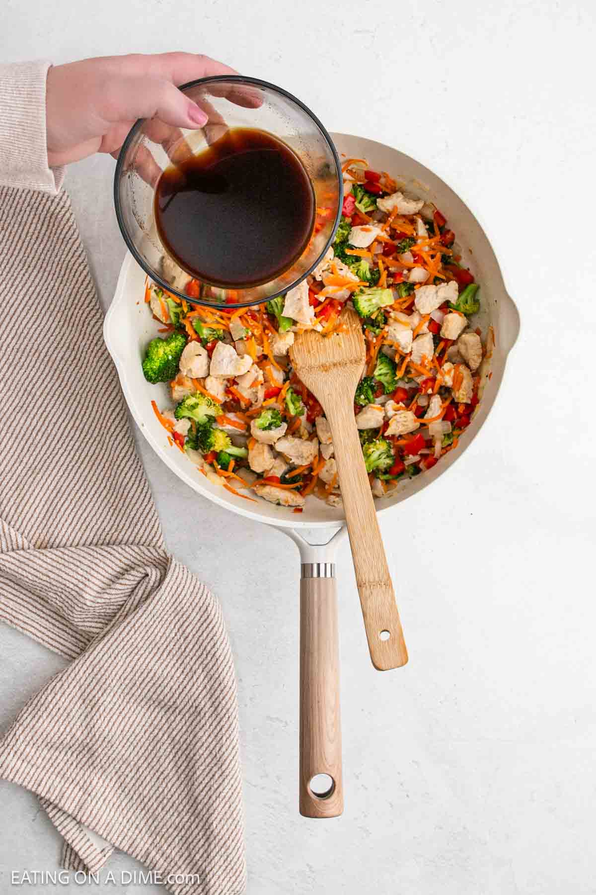 A hand pours dark sauce from a glass bowl into a white skillet filled with chicken, broccoli, carrots, and other vegetables—creating a Chicken Ramen-inspired dish. A wooden spoon rests in the skillet, with a beige cloth nearby on the white surface.