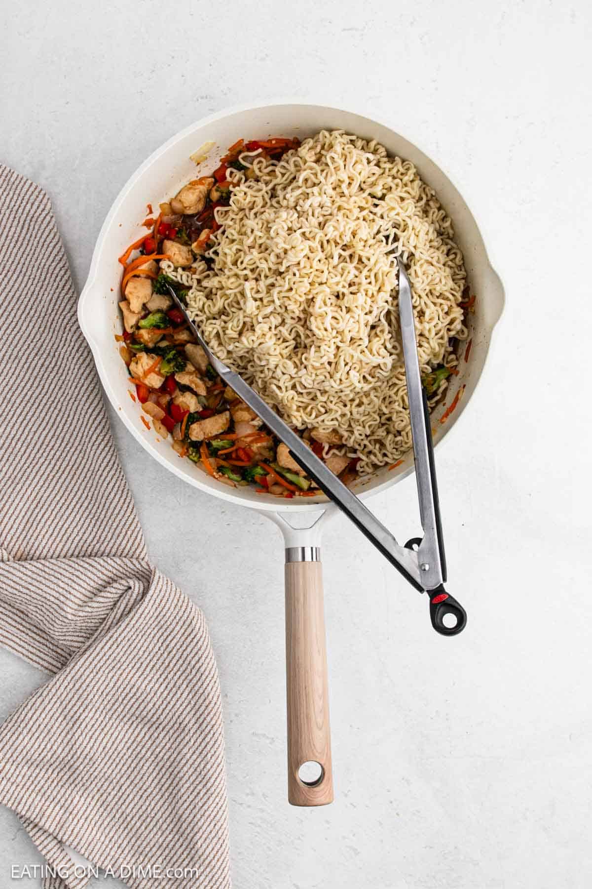 A white skillet filled with cooked chicken ramen noodles and mixed vegetables sits on a light surface with metal tongs resting on top. A beige striped kitchen towel is placed beside the skillet.