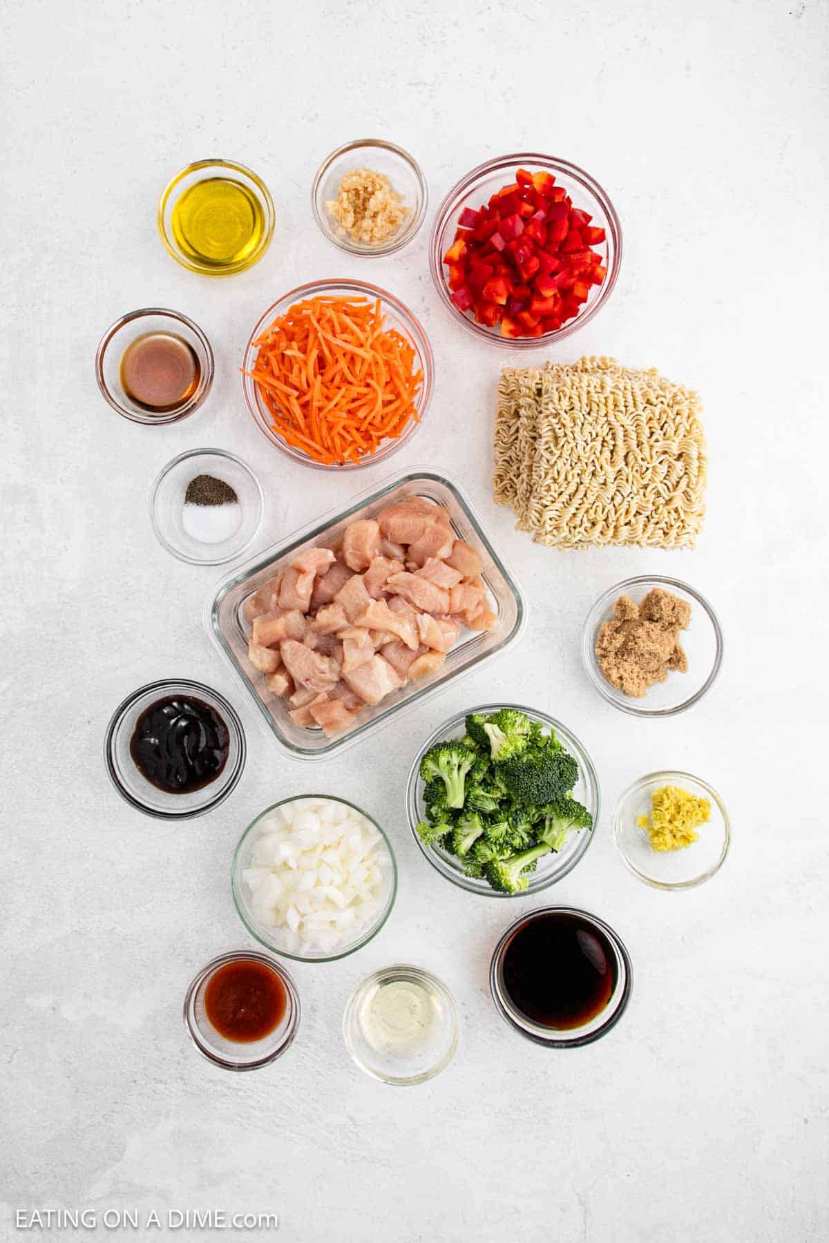 Overhead view of Chicken Ramen ingredients in glass bowls, including diced chicken, dry ramen noodles, chopped red bell pepper, broccoli, shredded carrots, onions, garlic, ginger, soy sauce, brown sugar, and oils on a white surface.