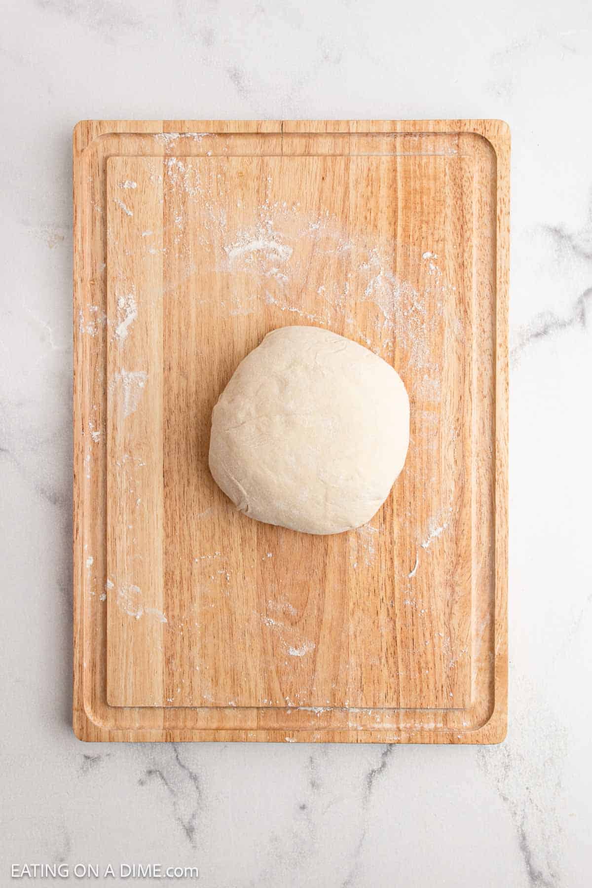 A ball of raw dough for sourdough bread sits on a lightly floured wooden cutting board, which rests on a white marble countertop.