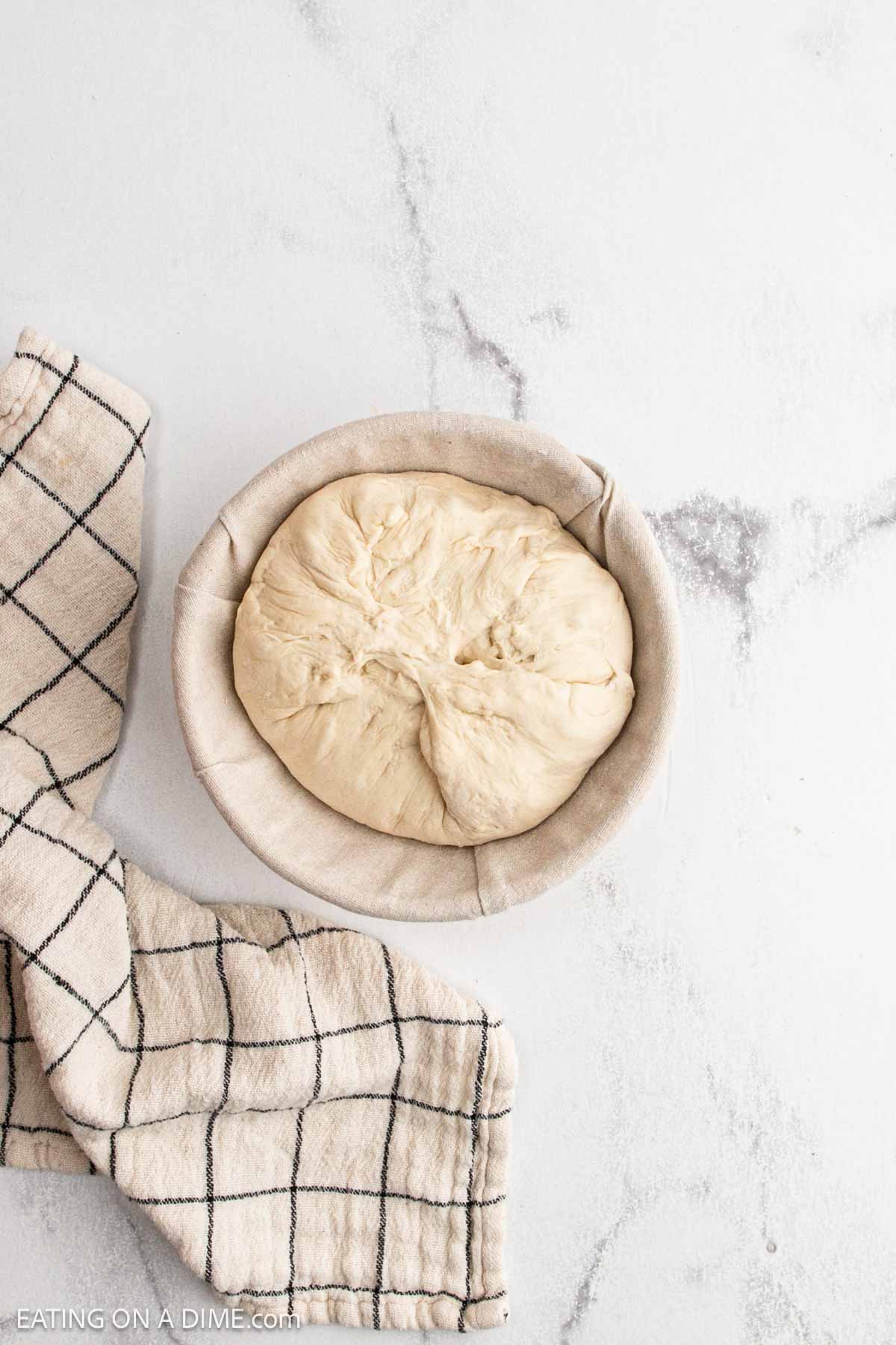 A round ball of bread dough rests in a cloth-lined bowl on a white surface, ready to make sourdough. A beige kitchen towel with a black grid pattern is placed beside it, setting the scene for crafting delicious sourdough bread.