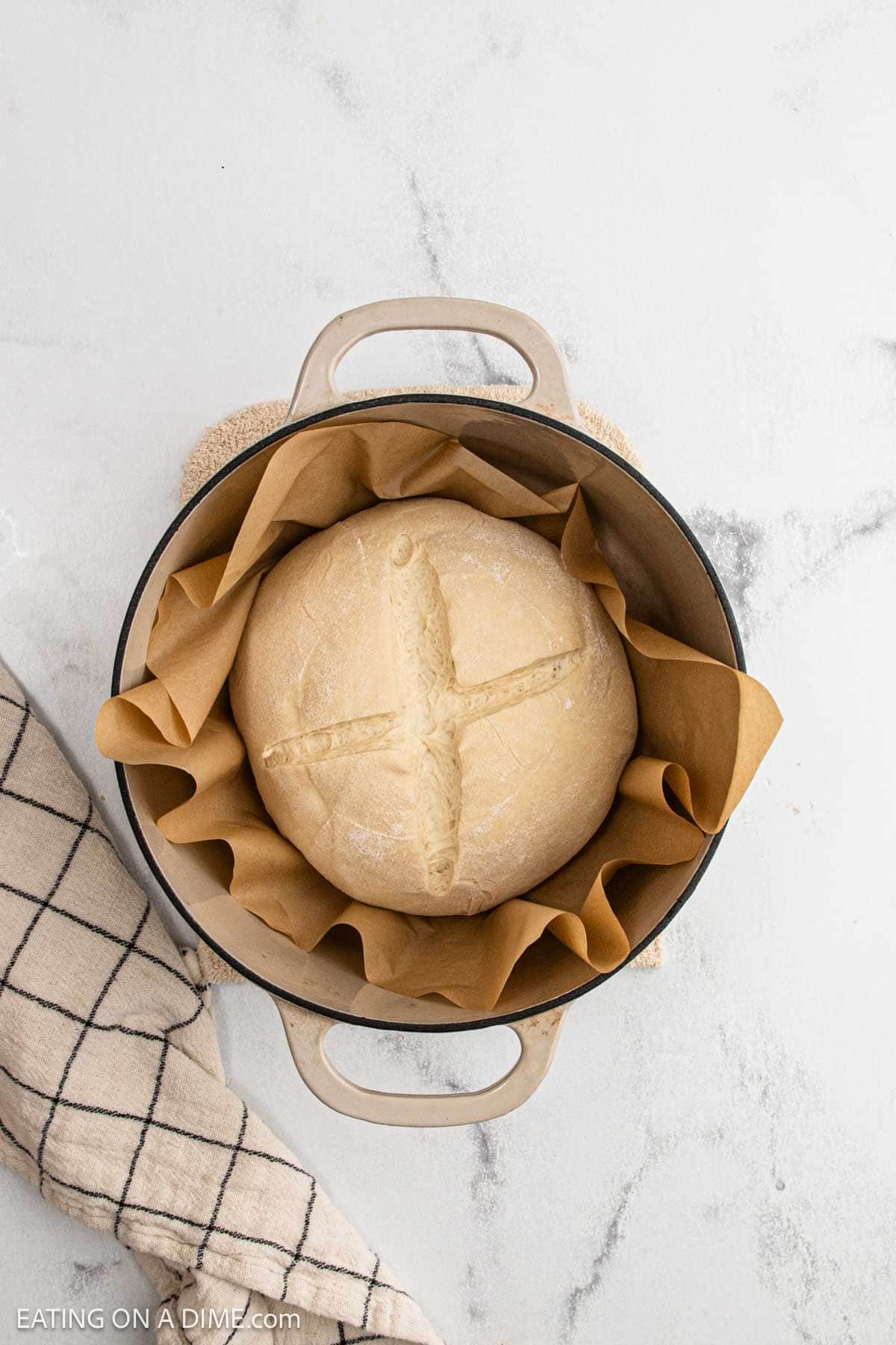 A round loaf of unbaked sourdough bread dough with a cross cut on top sits in a Dutch oven lined with brown parchment paper. A checkered towel lies nearby on a light-colored surface, ready to help you make sourdough at home.