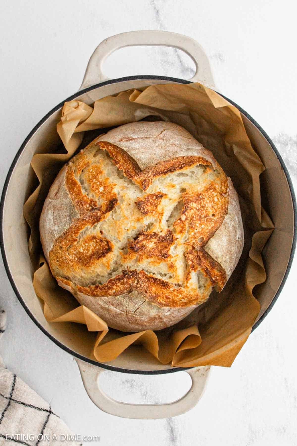 A round loaf of crusty artisan sourdough bread sits on brown parchment paper inside a white Dutch oven on a marble surface.