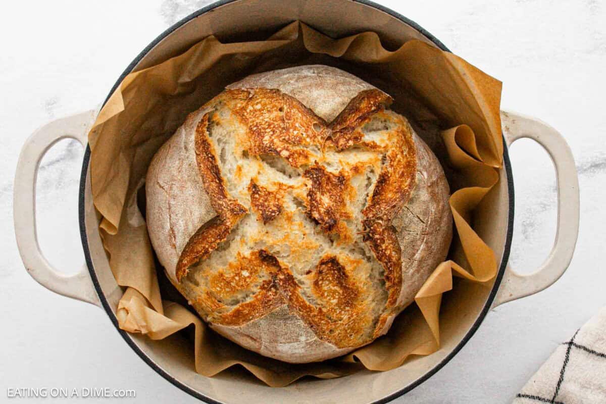 A round loaf of artisan sourdough bread with a golden, crusty top sits on parchment paper inside a white Dutch oven. The bread features a large, decorative X-shaped score on its surface.