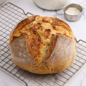 A round loaf of artisan sourdough bread with a golden, crusty top sits on a wire cooling rack. In the background, a metal measuring cup filled with flour and a glass jar hint at the sourdough recipe used to make this rustic treat.