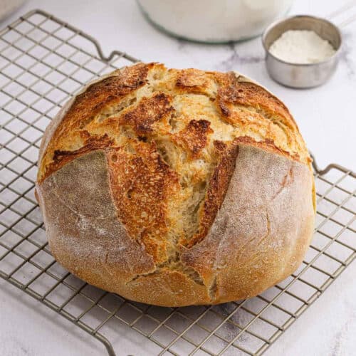 A round loaf of artisan sourdough bread with a golden, crusty top sits on a wire cooling rack. In the background, a metal measuring cup filled with flour and a glass jar hint at the sourdough recipe used to make this rustic treat.
