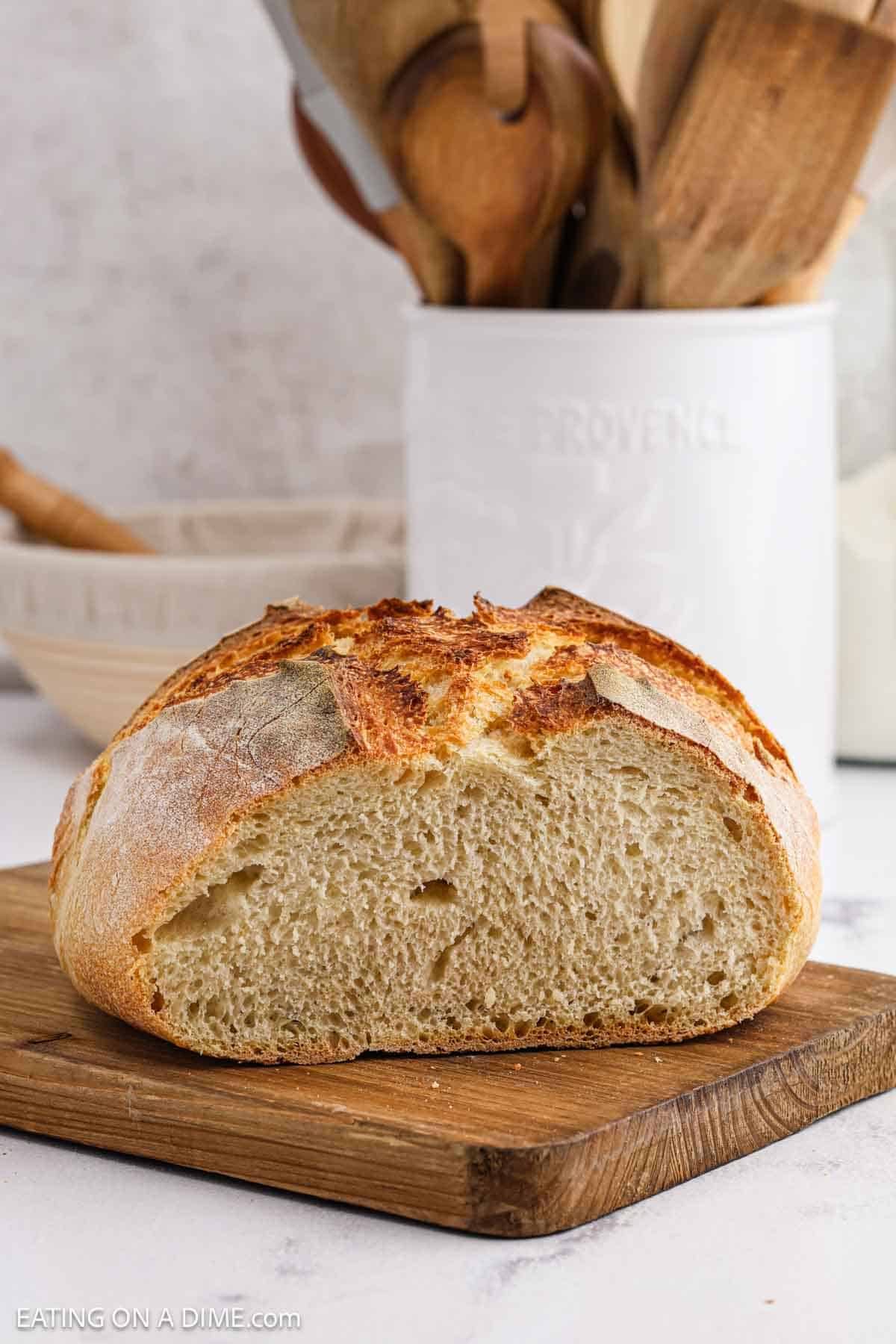 A round loaf of rustic sourdough bread, sliced in half to reveal its airy interior and golden, crusty exterior, sits on a wooden cutting board with kitchen utensils in the background.