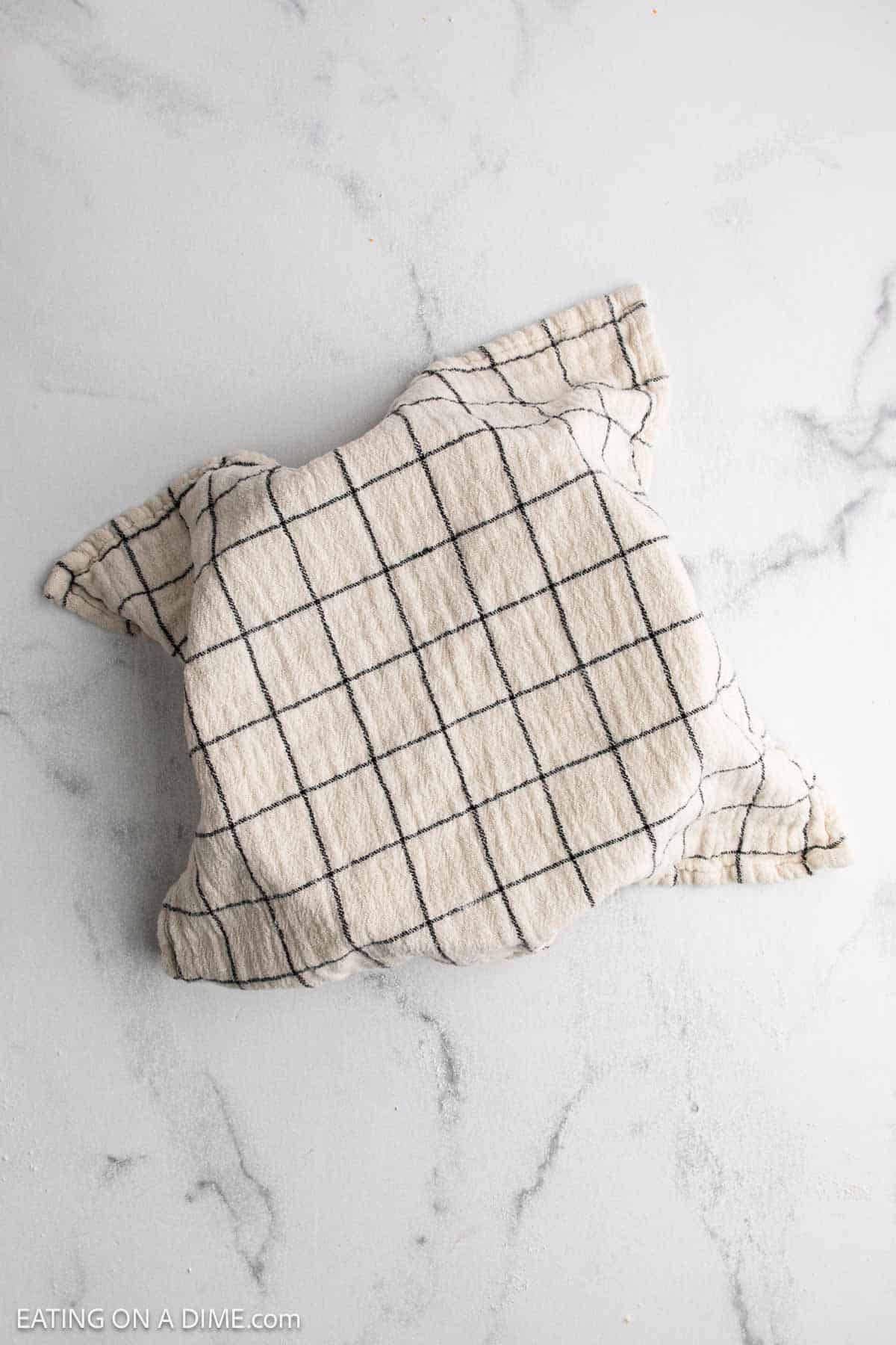 A beige kitchen towel with black grid lines covers a bowl on a light gray, marbled surface—perfect for letting your sourdough recipe rise as you make sourdough bread at home.