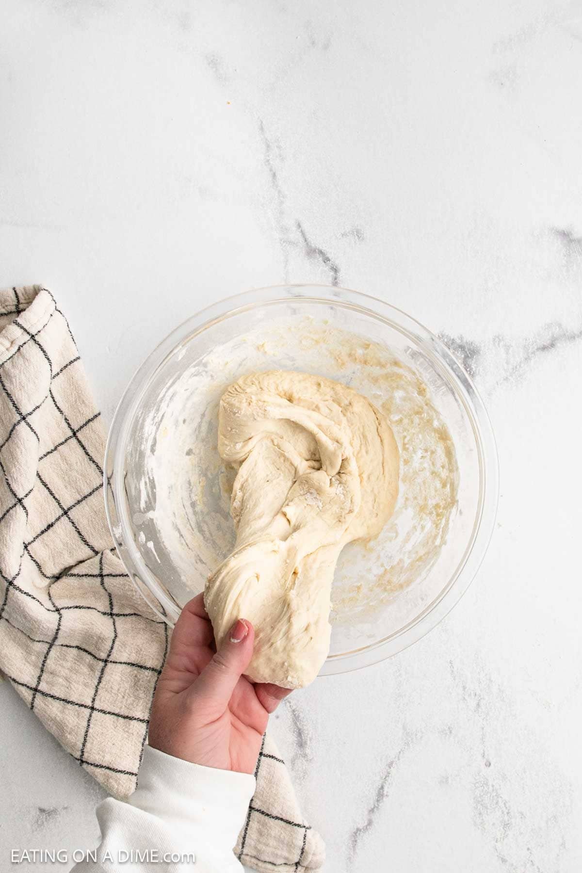 A hand holds stretchy sourdough bread dough above a glass mixing bowl on a white surface, with a beige and black checkered kitchen towel beside the bowl—perfect for anyone eager to make sourdough at home.