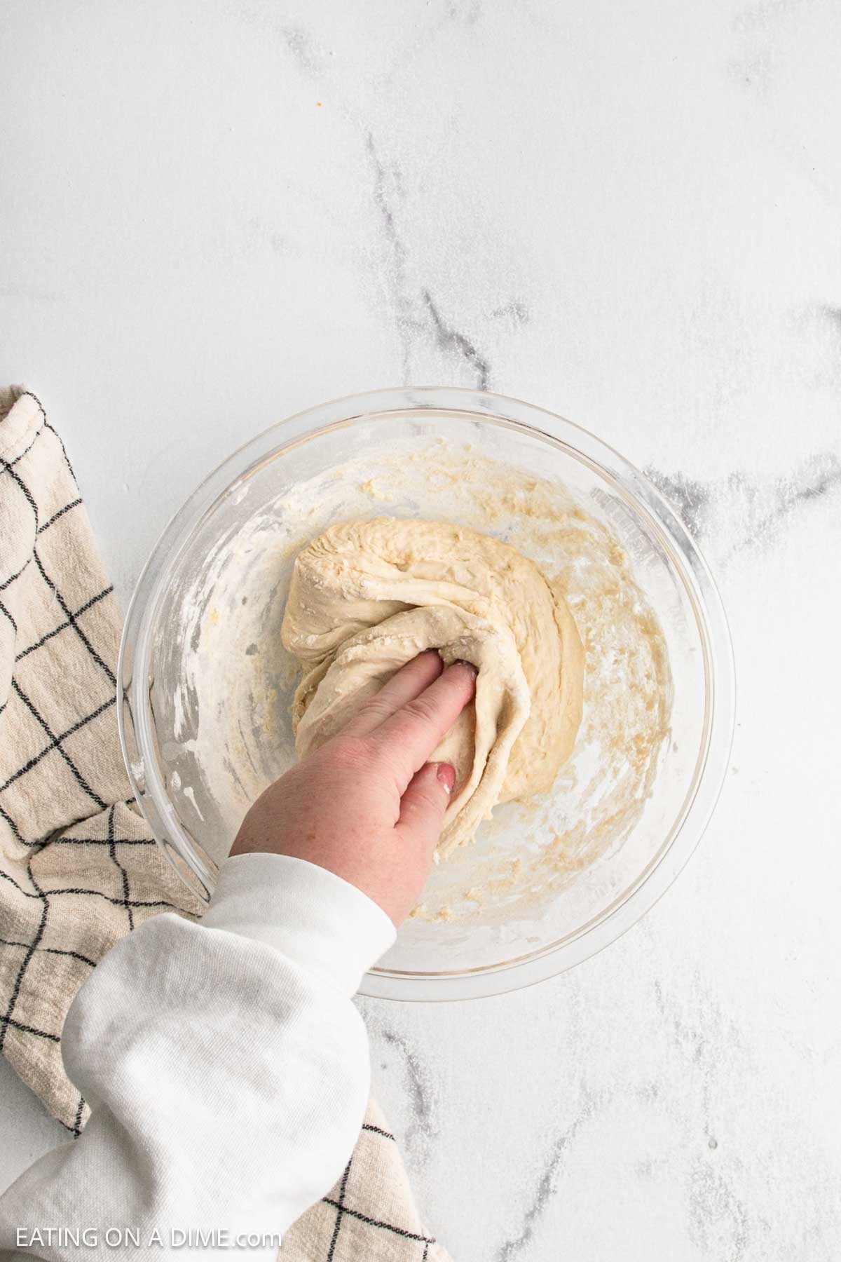 A hand kneads dough for sourdough bread in a clear glass bowl on a white surface, with a beige and black checkered towel nearby.
