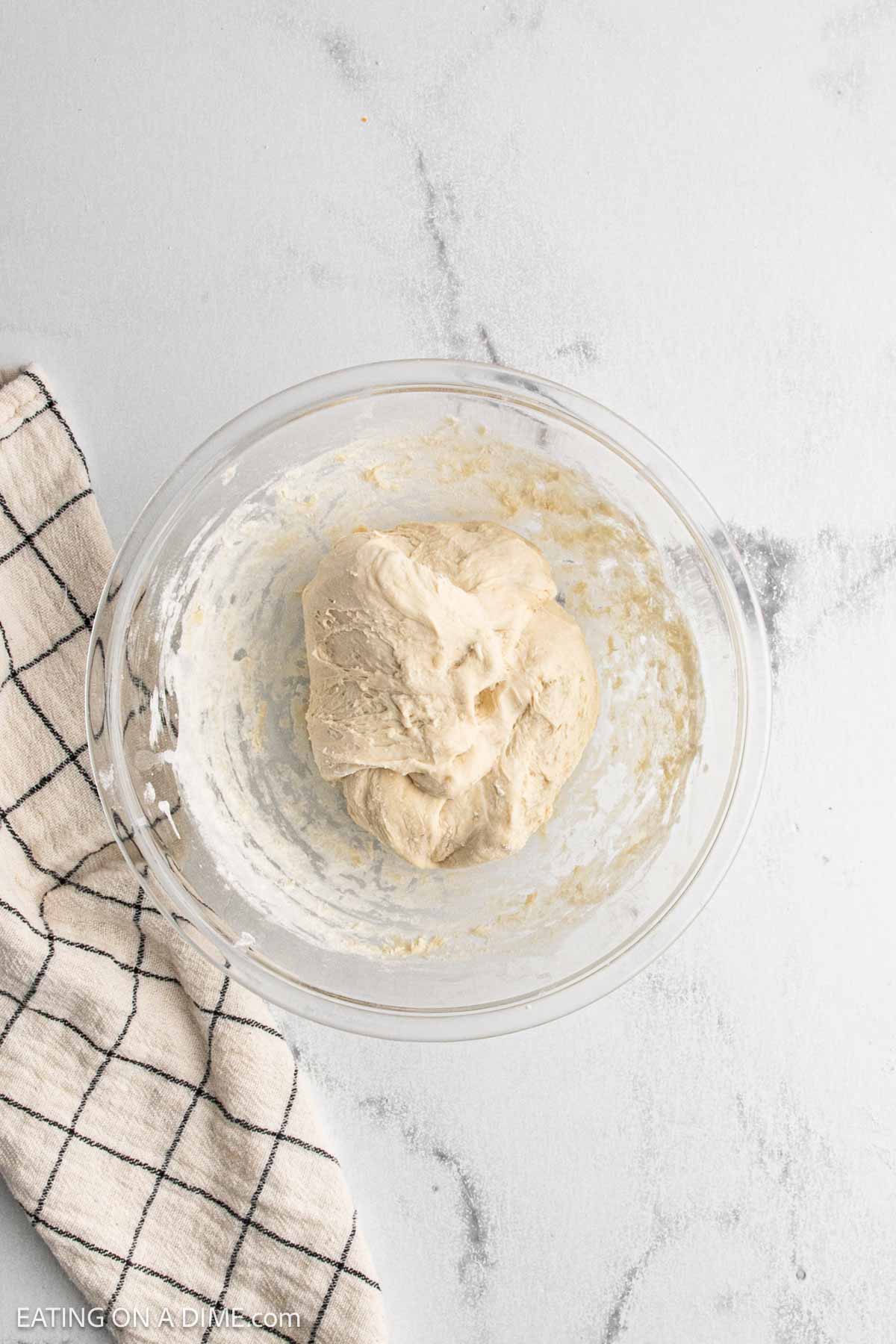 A glass bowl containing a lump of dough sits on a light gray countertop next to a beige kitchen towel with black grid lines, ready for your next sourdough bread recipe.
