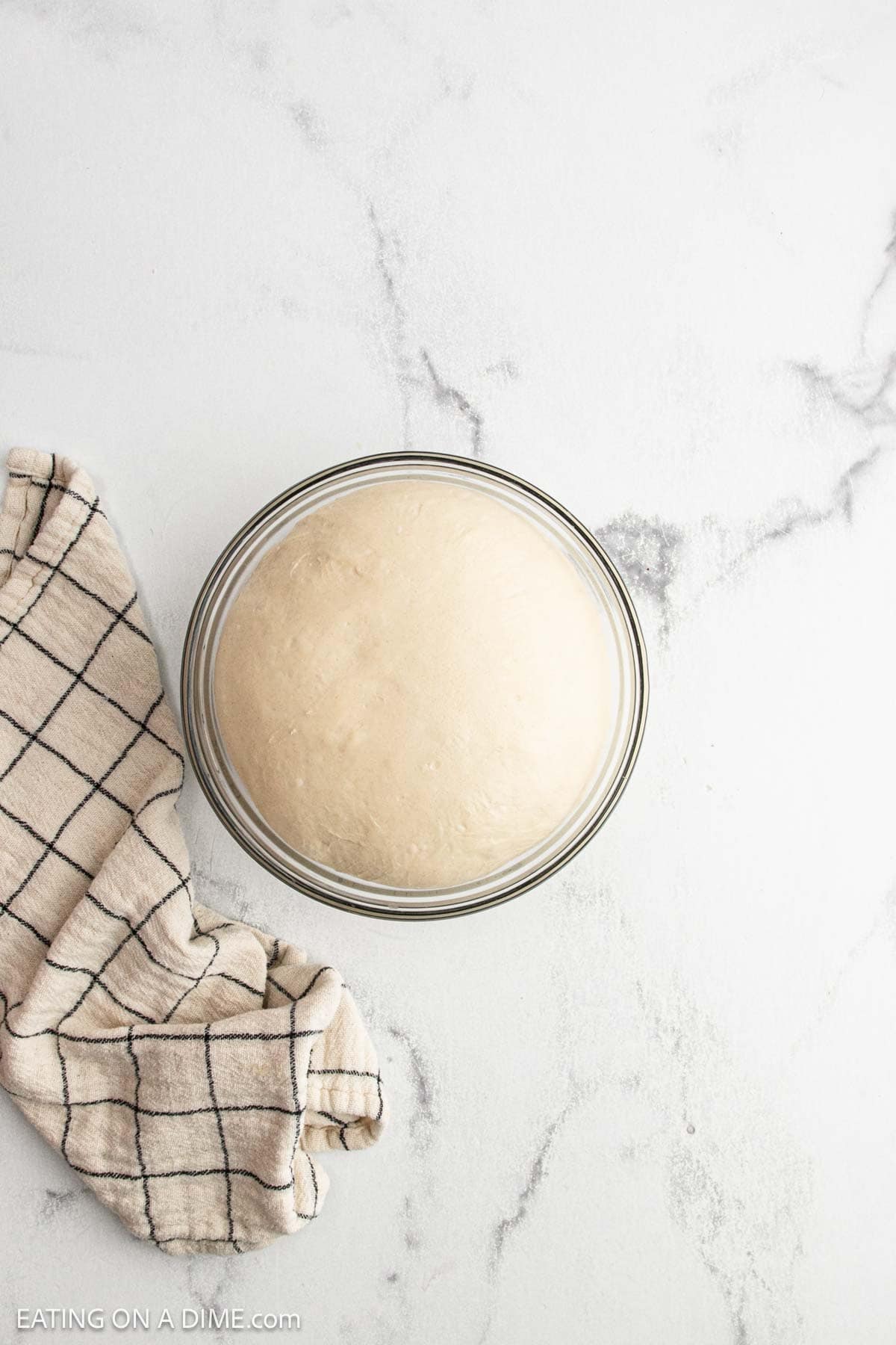 A glass bowl filled with risen sourdough bread dough sits on a white marble surface next to a beige and black checkered kitchen towel, ready for the next step in your sourdough bread recipe.