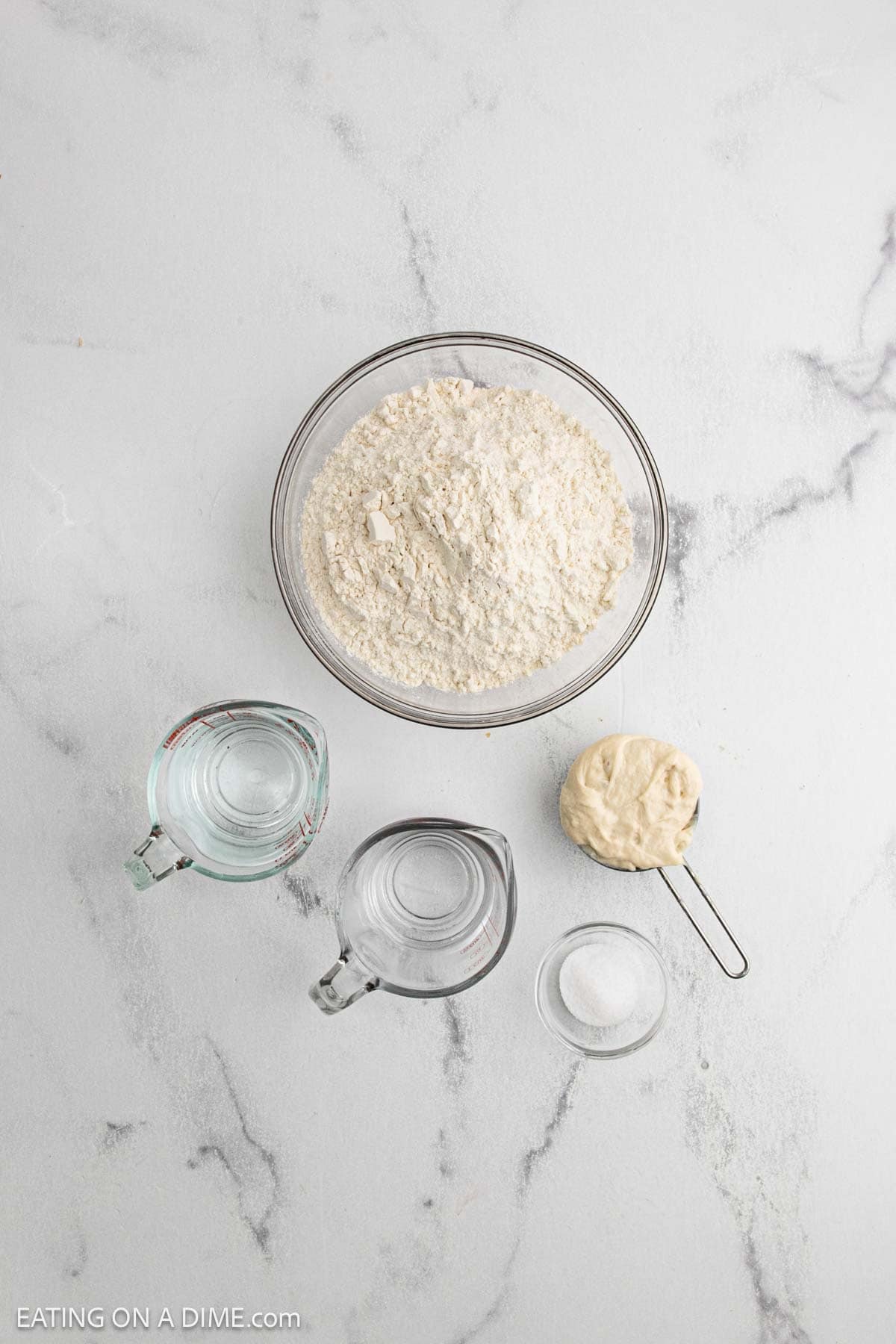 A glass bowl of flour, a measuring cup of dough, two glass measuring cups with water, and a small bowl of salt are arranged on a white marble surface—simple essentials to make sourdough or follow your favorite sourdough recipe.