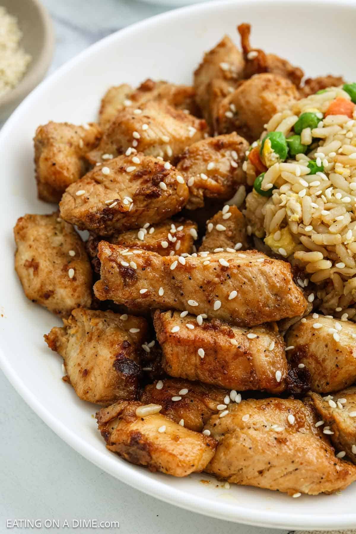 A white bowl filled with browned, seasoned Chicken pieces sprinkled with sesame seeds, served next to vegetable fried rice with peas and carrots.