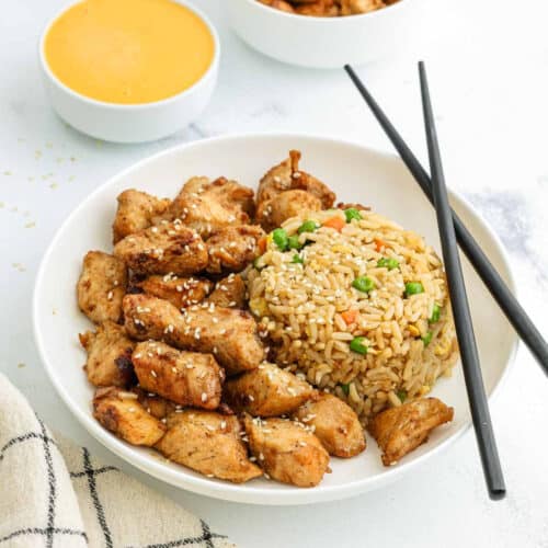 A white plate with Hibachi Chicken pieces sprinkled with sesame seeds, a mound of fried rice with peas and carrots, and black chopsticks. In the background, there is a bowl of yellow dipping sauce.