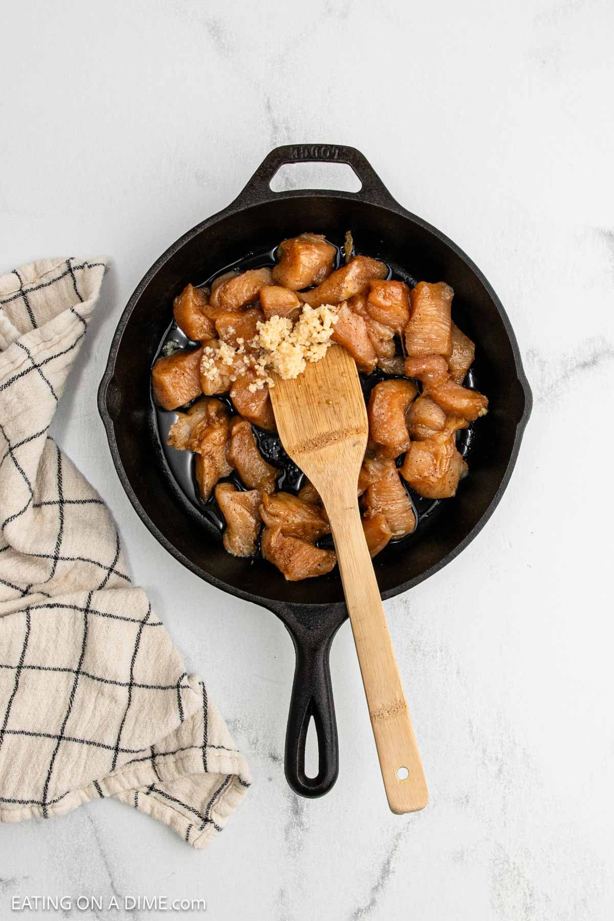 A cast iron skillet with chunks of seasoned Chicken and minced garlic being stirred with a wooden spatula,