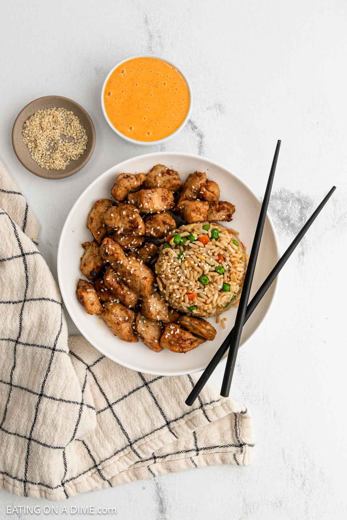 Golden brown chicken pieces on a white with a serving of fried rice with peas and carrots, black chopsticks, a bowl of orange dipping sauce, a small bowl of sesame seeds, and a checkered cloth—classic Japanese cuisine at its finest.