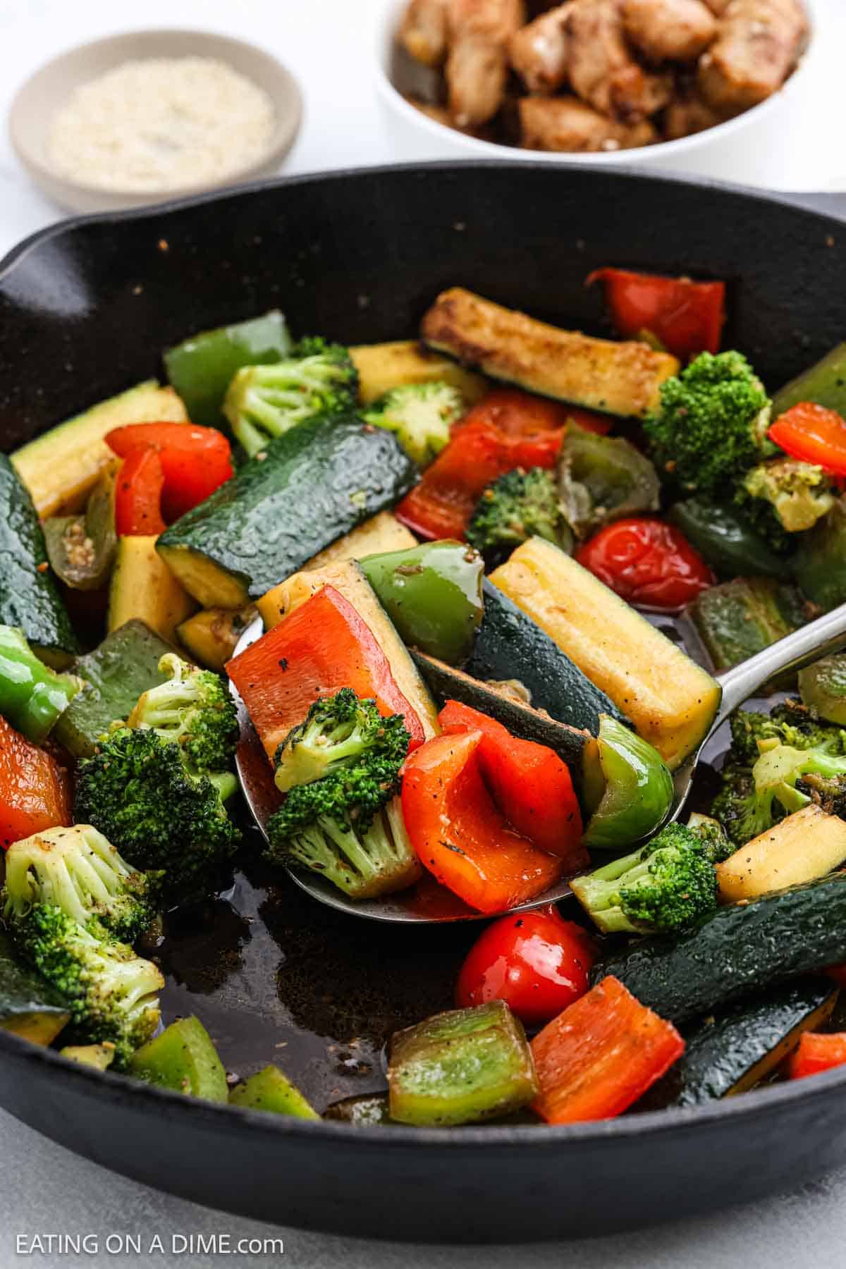 A close-up of a skillet filled with colorful Hibachi Vegetables, like broccoli, zucchini, red and green bell peppers. A spoon lifts a portion while bowls of chicken and sesame seeds are in the background.