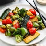 A plate of hibachi vegetables, including stir-fried broccoli, red and green bell peppers, and zucchini, is garnished with sesame seeds. Black chopsticks rest on the plate's edge.