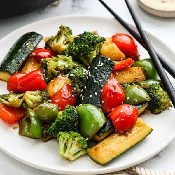 A plate of hibachi vegetables, including stir-fried broccoli, red and green bell peppers, and zucchini, is garnished with sesame seeds. Black chopsticks rest on the plate's edge.