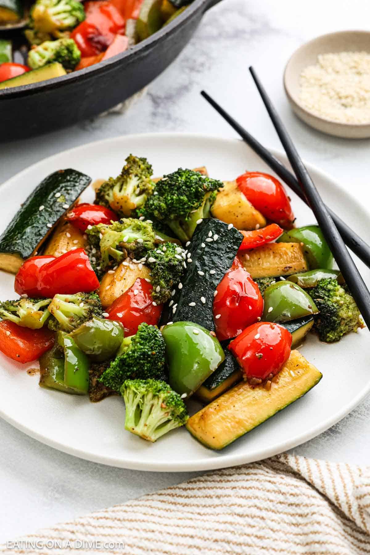 A plate of hibachi vegetables, including broccoli, zucchini, red bell pepper, and green bell pepper, garnished with sesame seeds. Black chopsticks rest on the side, and a small dish of sesame seeds is visible in the background.