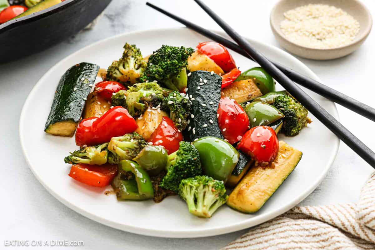 A white plate with hibachi vegetables including broccoli, zucchini, red and green bell peppers, and cherry tomatoes, garnished with sesame seeds. Black chopsticks and a small bowl of sesame seeds are next to the plate.