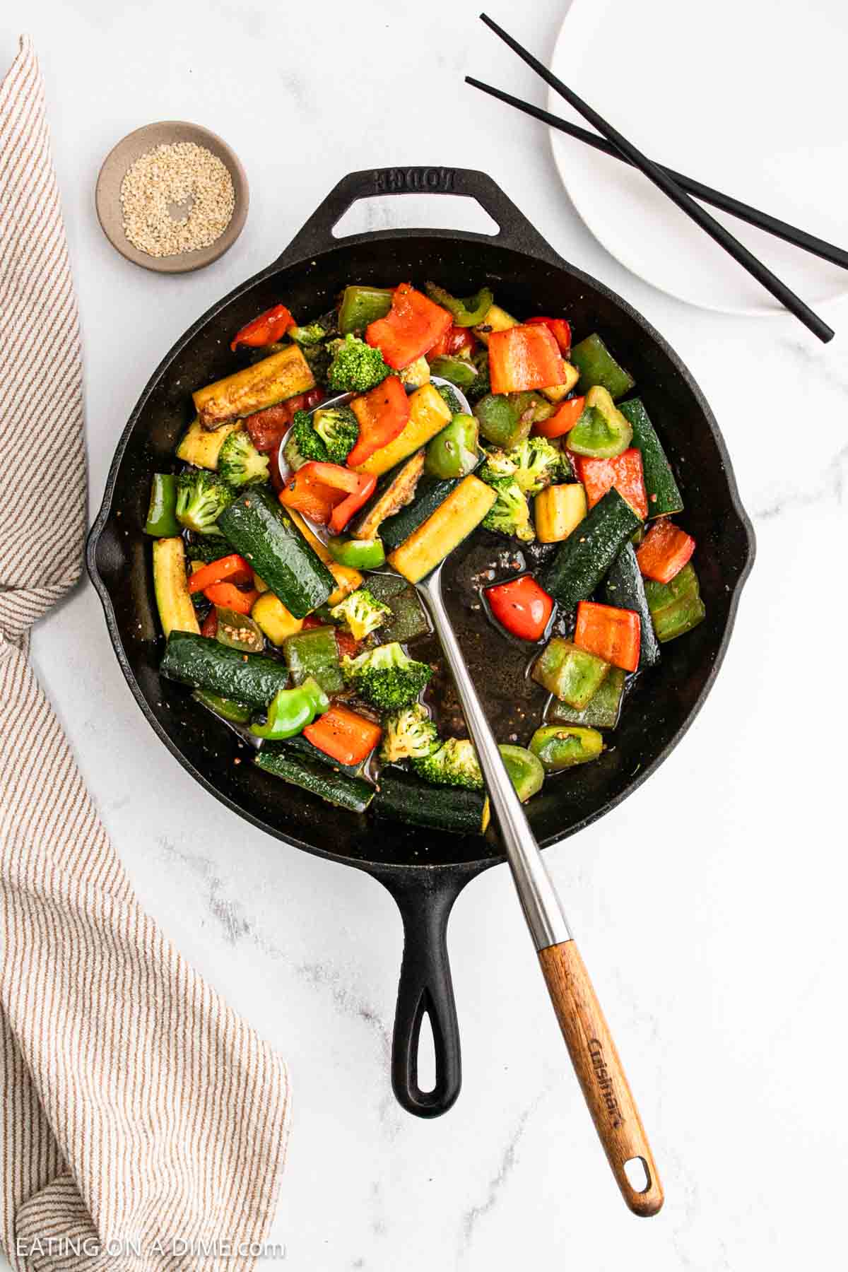A cast iron skillet filled with Hibachi Vegetables—broccoli, bell peppers, and zucchini—sits on a white surface with a striped towel, chopsticks, a small dish of sesame seeds, and a white plate nearby.