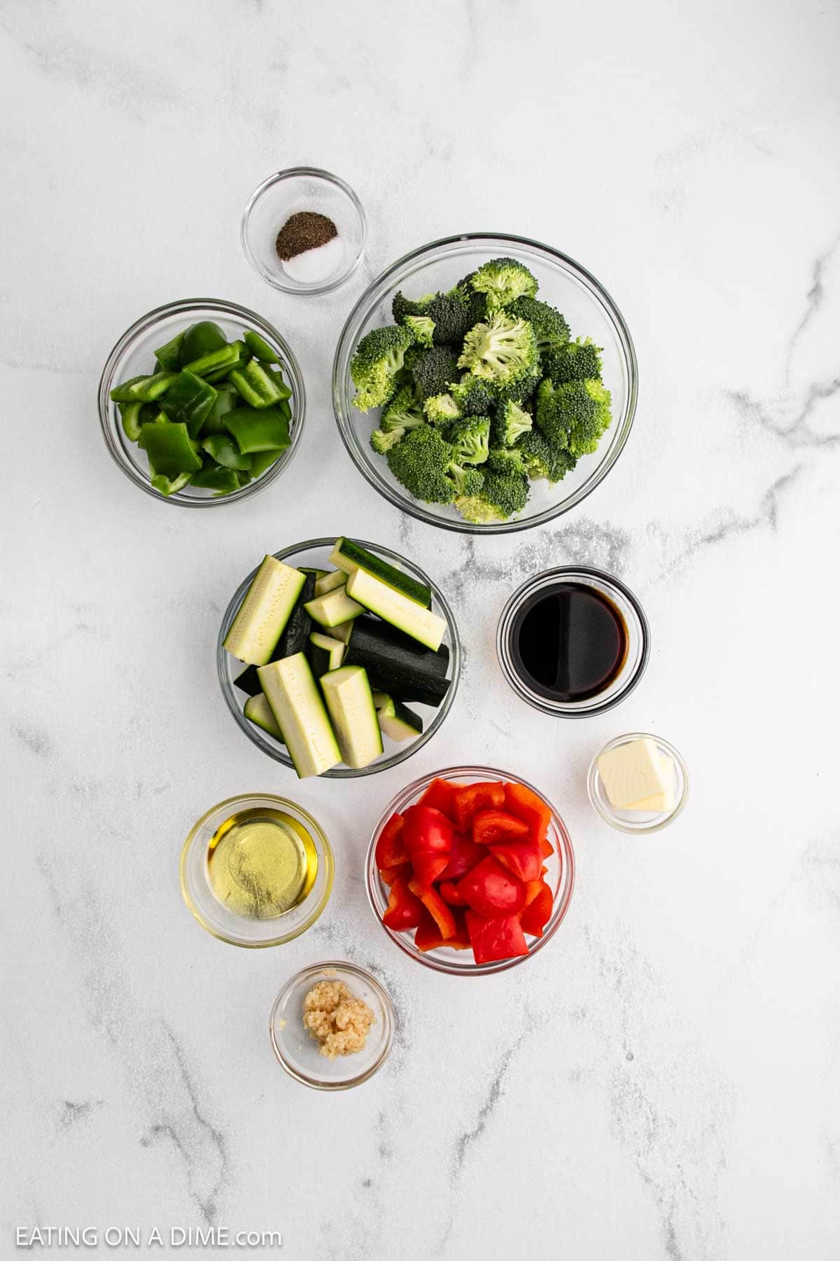 Overhead view of bowls containing Hibachi Vegetables like green bell pepper, broccoli florets, zucchini sticks, red bell pepper, soy sauce, butter, olive oil, minced garlic, and black pepper on a white marble surface.