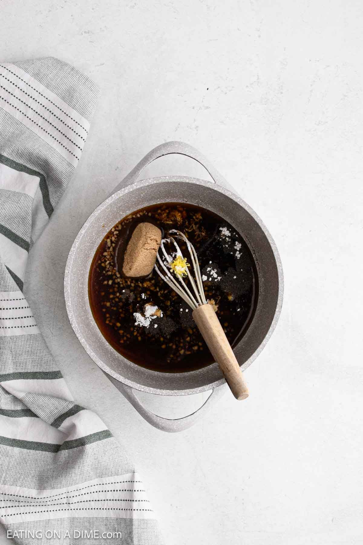 A gray saucepan filled with sauce ingredients, like brown sugar, minced garlic, and seasonings, sits on a white surface next to a gray striped kitchen towel. A whisk rests in the pot.