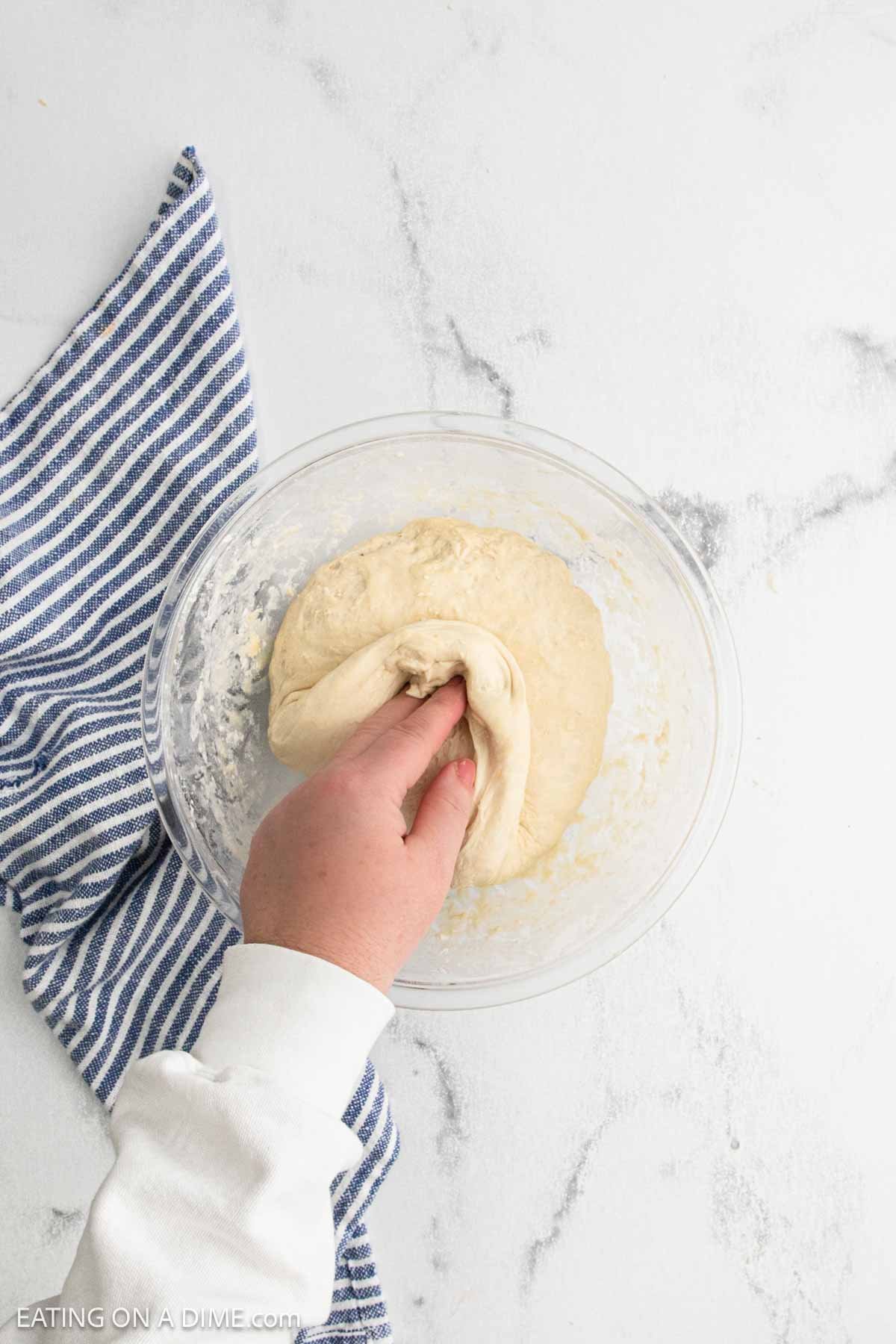 A hand kneading sourdough bread dough in a clear glass bowl on a white surface, next to a blue and white striped kitchen towel.