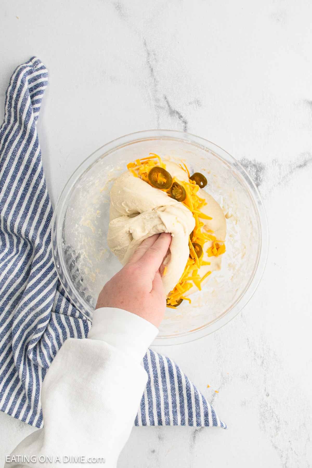 A hand kneads sourdough bread dough in a glass bowl with shredded cheddar cheese and jalapeño slices on a white surface, next to a blue and white striped kitchen towel.