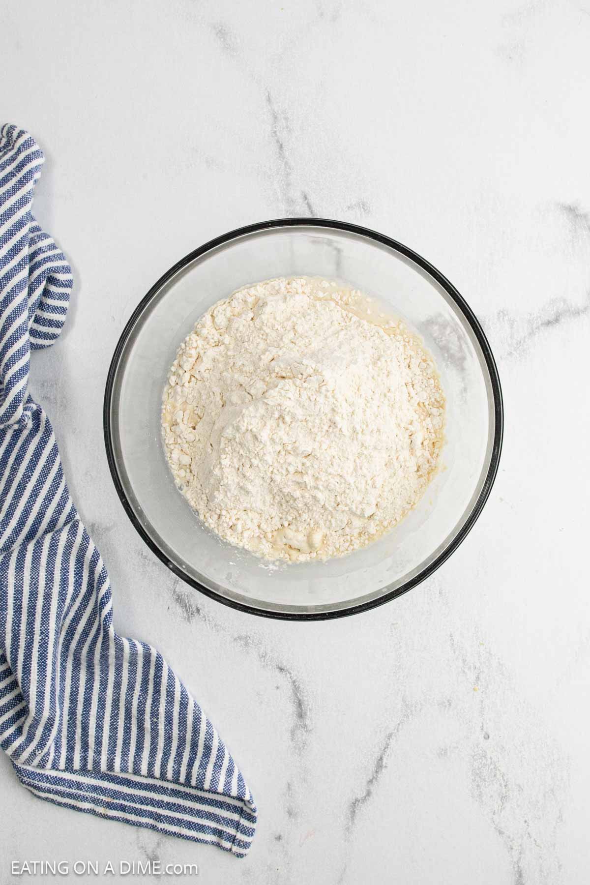 A glass bowl filled with flour mixture for sourdough bread sits on a white marble surface next to a blue and white striped kitchen towel.