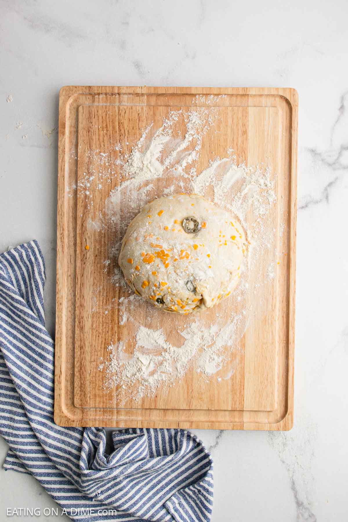 A round ball of sourdough bread dough, studded with cheddar and jalapeño, sits on a floured wooden cutting board. A blue and white striped cloth is placed nearby on a white marble surface.
