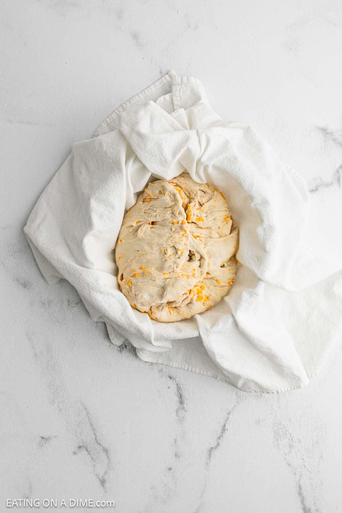 A round loaf of unbaked Cheddar Sourdough bread dough with small orange specks is wrapped in a white cloth on a light marble surface.