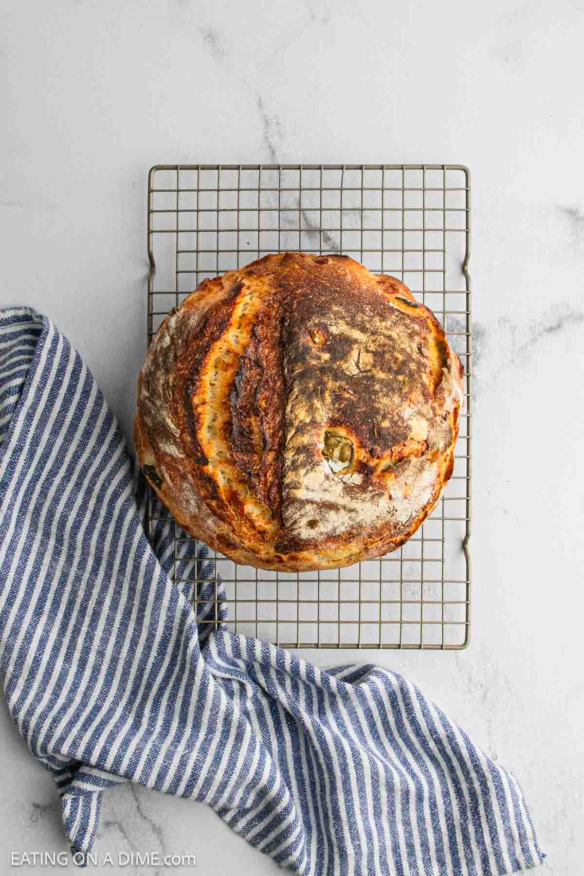 A round loaf of rustic sourdough bread with a golden-brown crust sits on a cooling rack, next to a blue and white striped kitchen towel on a light marble surface.