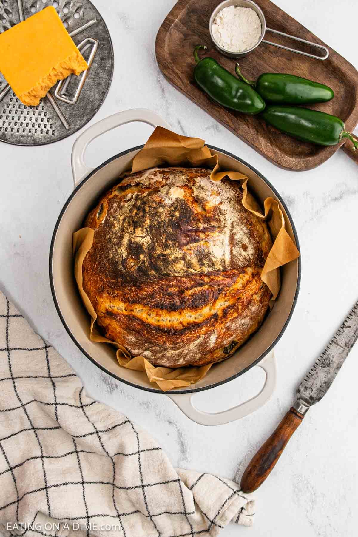 A round loaf of crusty jalapeño cheddar sourdough bread in a Dutch oven with parchment paper, surrounded by a block of cheddar cheese, two jalapeños, a cup of flour, a grater, cloth, and bread knife on a light surface.