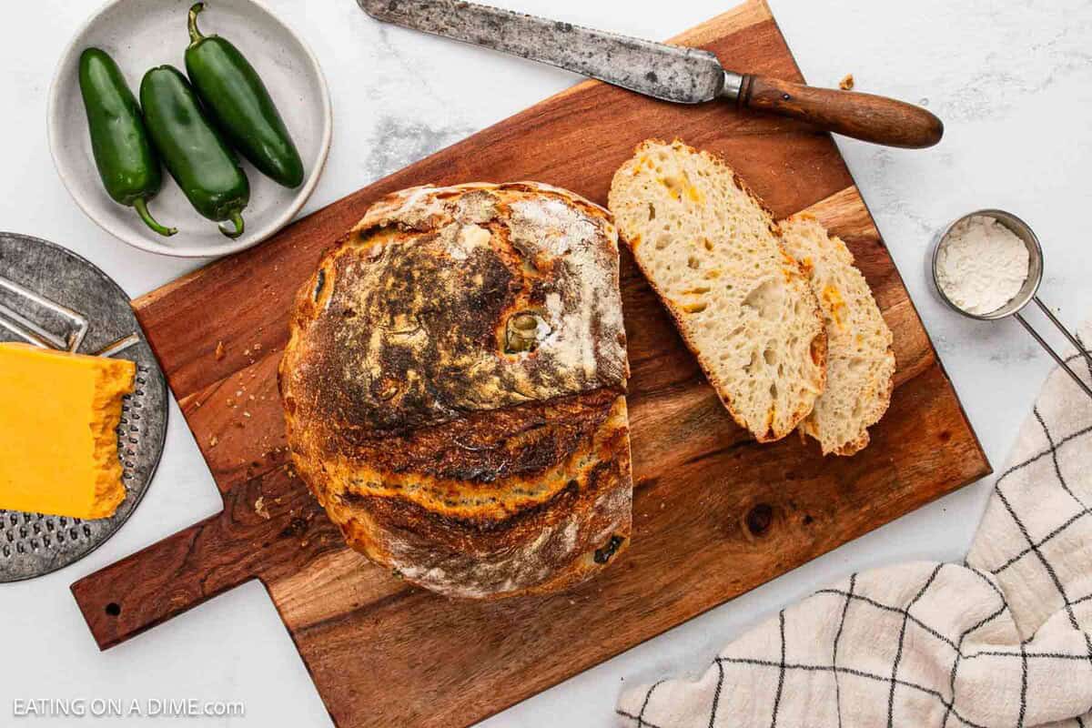 A loaf of rustic sourdough bread with two slices cut sits on a wooden cutting board next to a bread knife, cheddar cheese, jalapeño peppers, a small bowl of flour, and a kitchen towel.