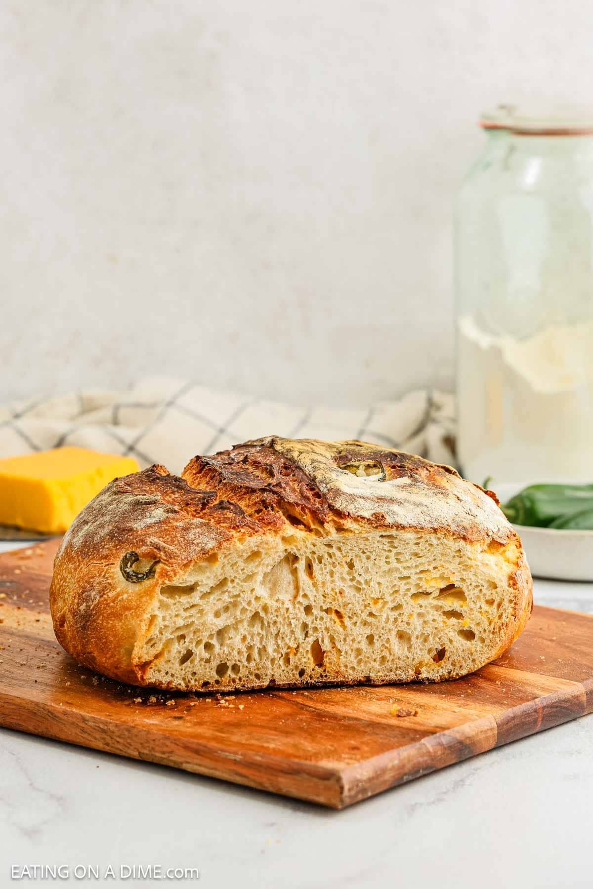 A rustic jalapeño sourdough bread with a golden crust sits on a wooden cutting board. The airy loaf, dotted with visible holes, is complemented by a block of cheddar, a jar of flour, and a cloth in the background.