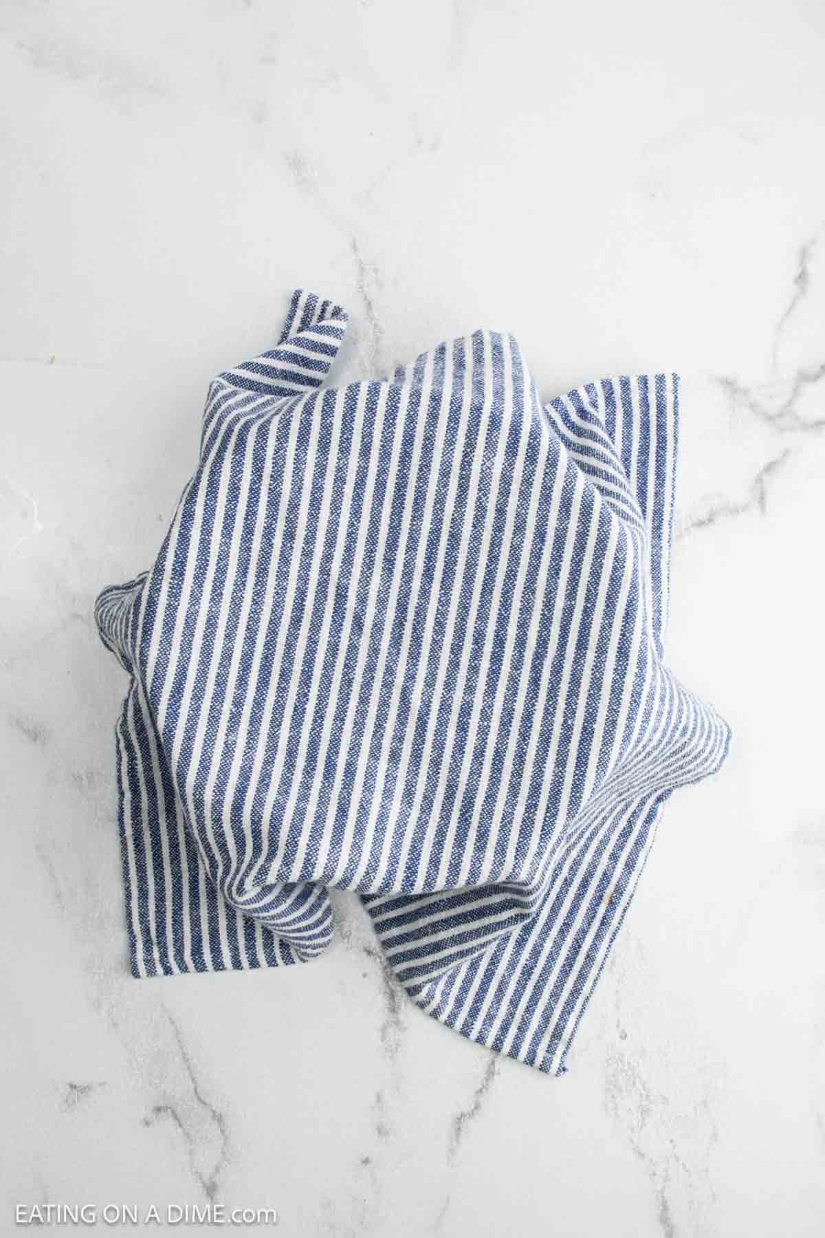 A blue and white striped cloth covers a round cheddar bread on a white marble surface. The edges of the cloth are loosely draped around the sourdough loaf.