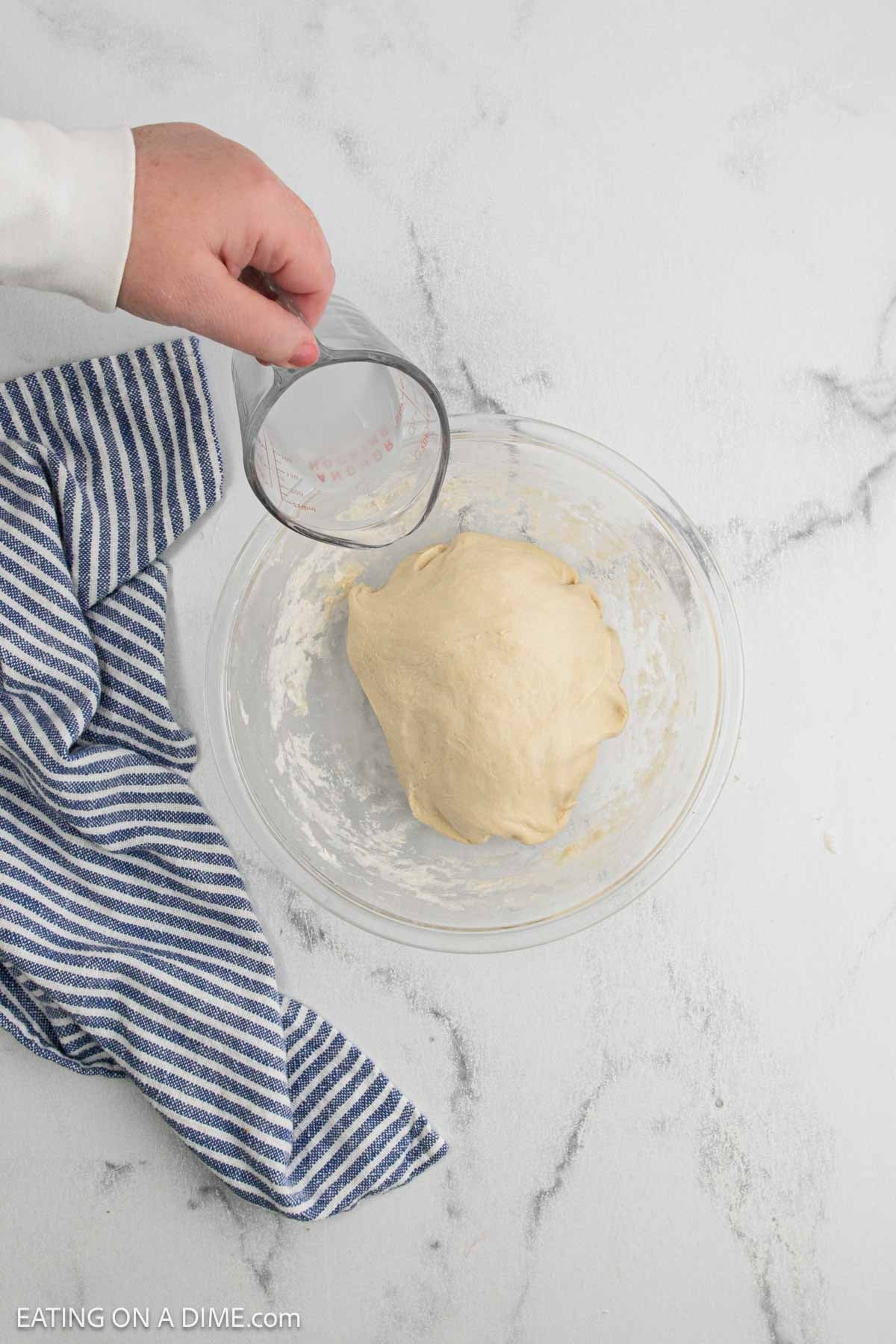 A hand holds a glass cup over a bowl of sourdough bread dough on a white surface. A blue and white striped towel lies beside the bowl.