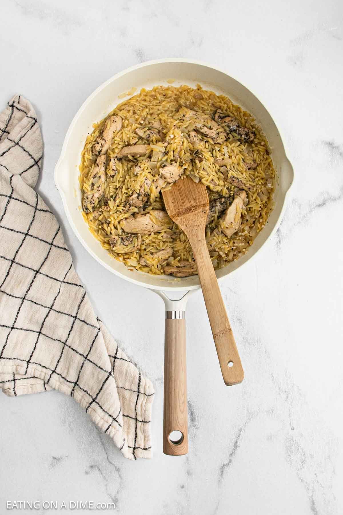 A skillet filled with Lemon Chicken Orzo and pieces of chicken, with a wooden spoon resting inside. A checkered kitchen towel is placed beside the skillet on a white marble surface.