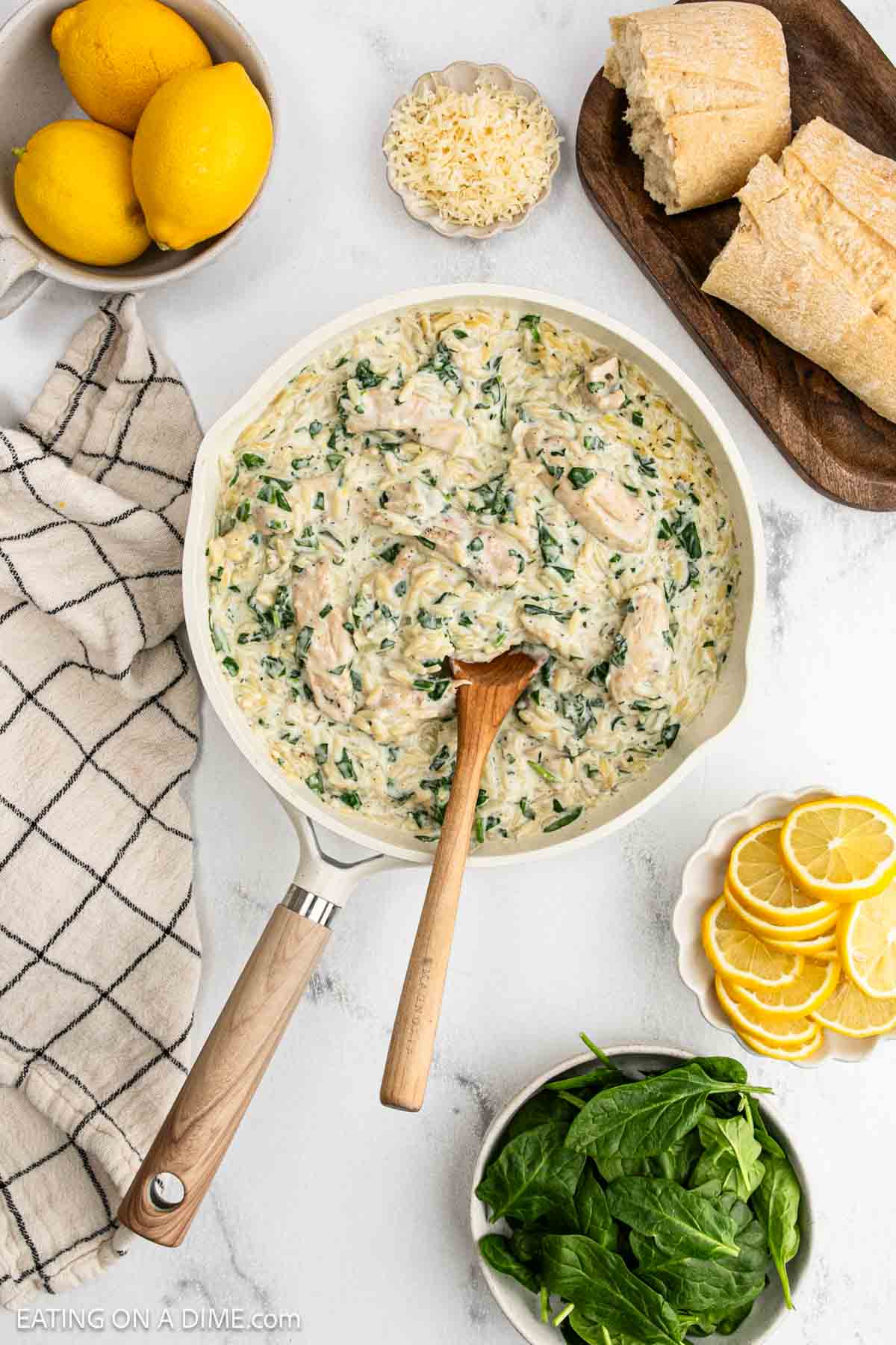 A skillet filled with creamy Lemon Chicken Orzo and spinach pasta, stirred with a wooden spoon, is surrounded by fresh lemons, grated cheese, a loaf of bread, lemon slices, baby spinach, and a checked towel on a white background.