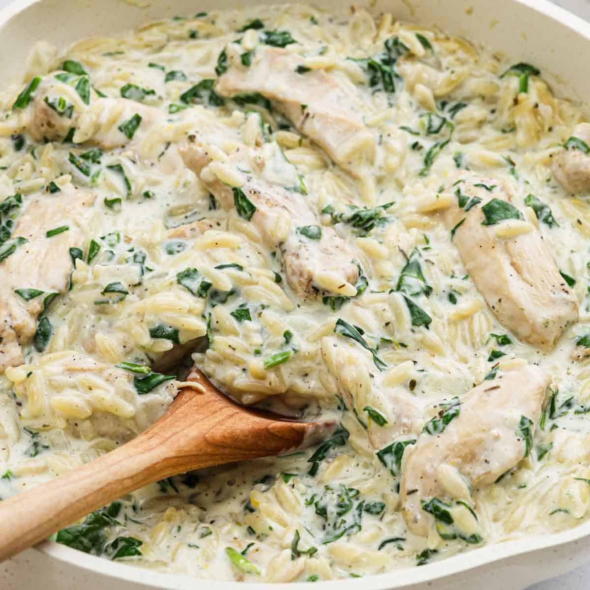 A creamy Lemon Chicken Orzo dish with spinach and tender chicken pieces, all mixed together in a thick white sauce, being stirred with a wooden spoon.