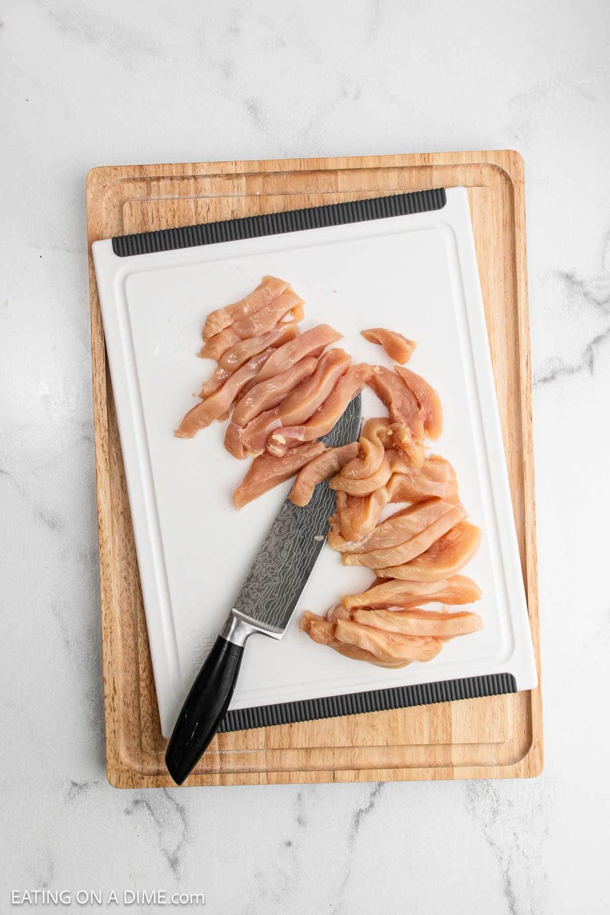 Raw chicken breast cut into strips on a white cutting board with a knife, ready for Lemon Chicken Orzo, placed on a wooden board atop a white marble surface. The text "EATINGONA DIME.com" appears in the bottom left corner.
