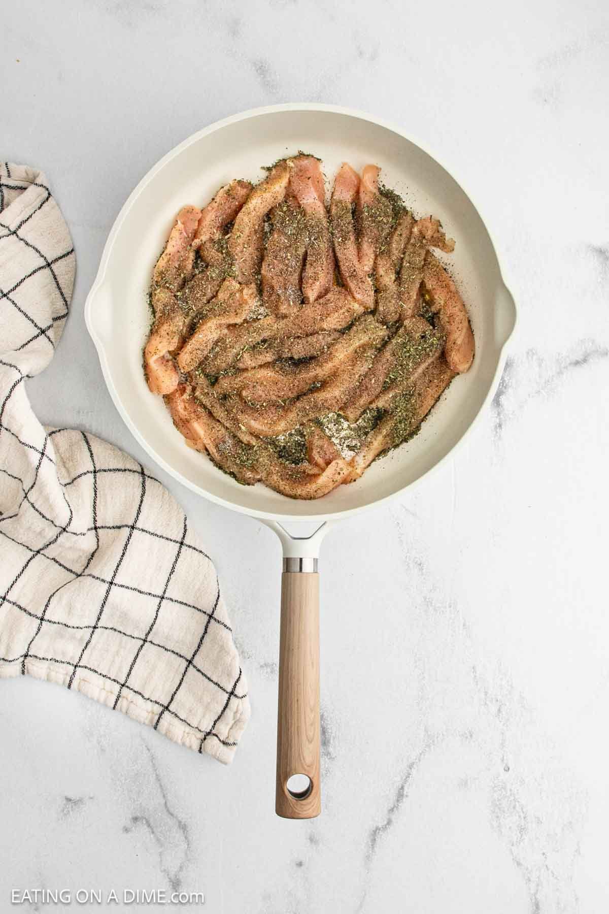 A white skillet filled with raw, seasoned strips of chicken for Lemon Chicken Orzo sits on a white marble surface next to a beige and black checkered kitchen towel.