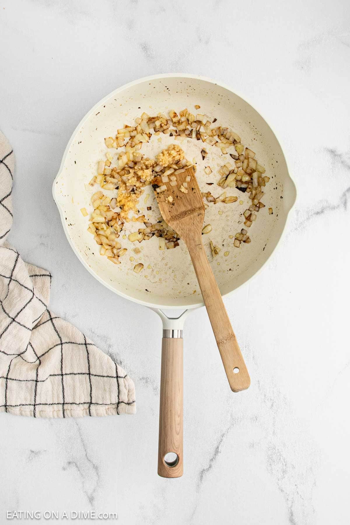 A white skillet with sautéed diced onions and minced garlic, stirred with a wooden spatula, sits on a white marble surface—perfect for starting a Lemon Chicken Orzo recipe—next to a beige and black checkered kitchen towel.