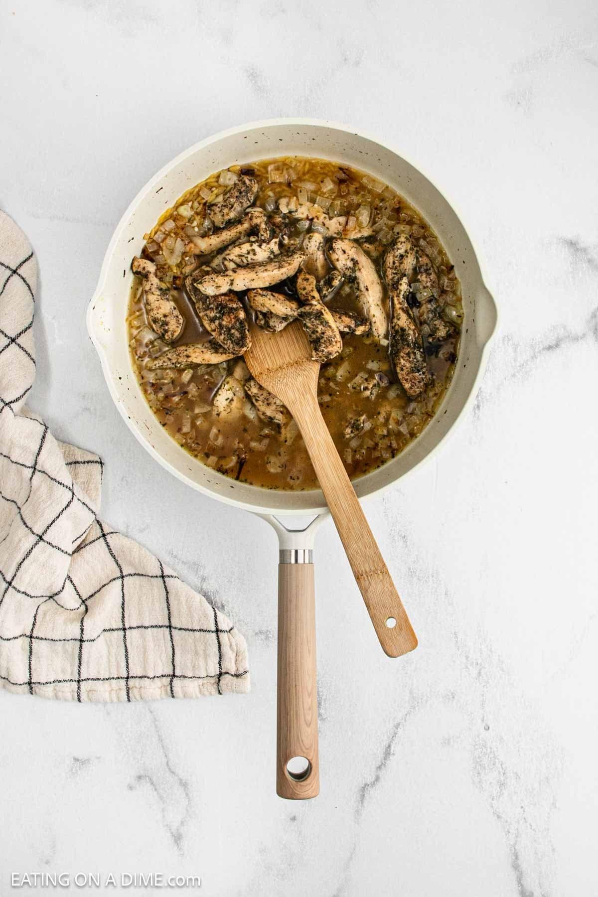 A skillet with cooked seasoned chicken strips and onions in broth for Lemon Chicken Orzo, stirred with a wooden spatula, sits on a white marble surface next to a checkered kitchen towel.