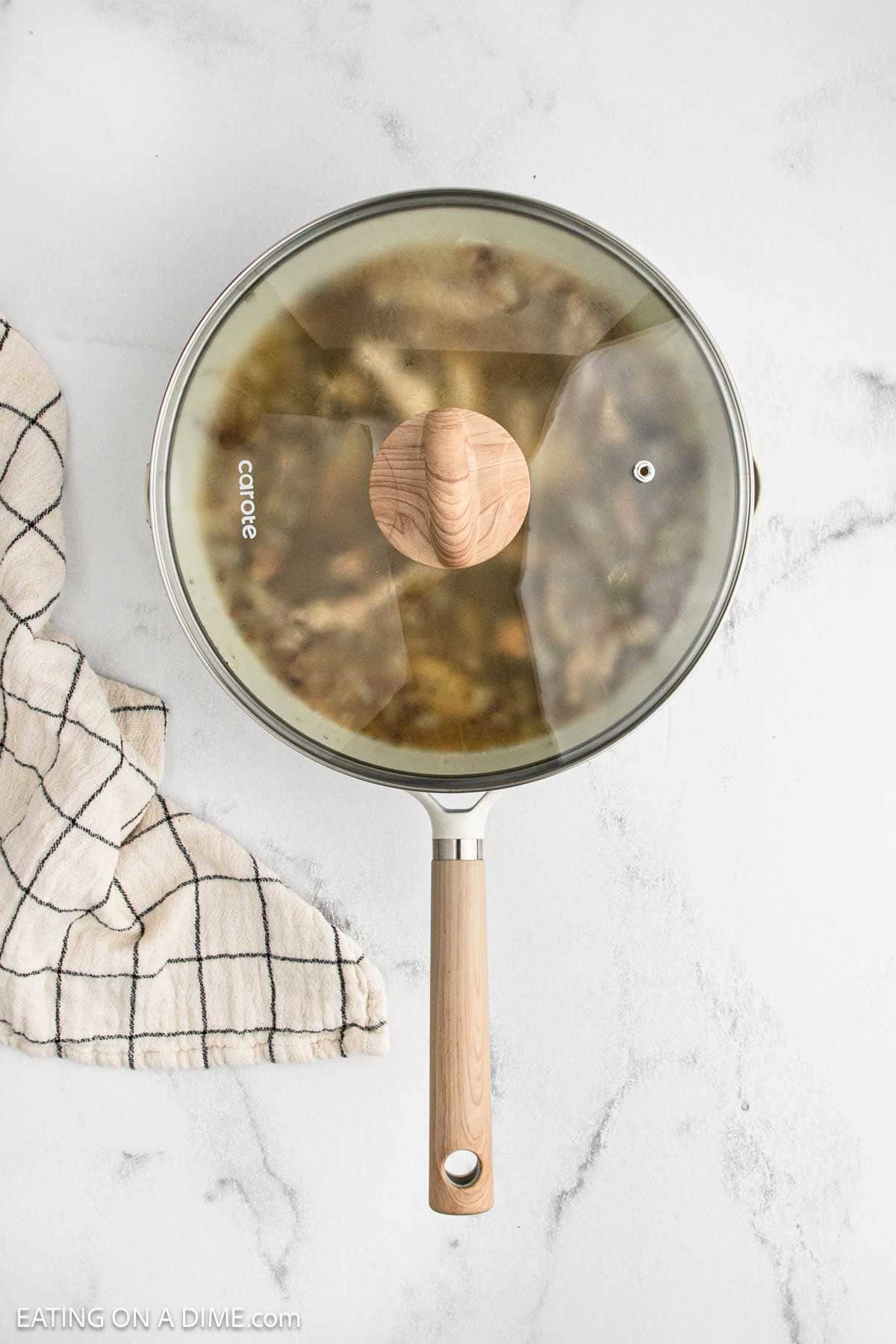 A frying pan with a glass lid and wooden handle sits on a white surface next to a beige and black checkered kitchen towel. Inside, savory Lemon Chicken Orzo simmers, visible through the lid.